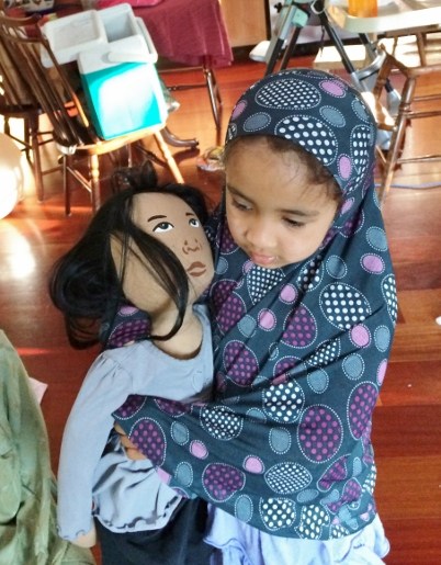 A child wearing a hijab with circles of different colors and sizes that have polka dots on the inside hugging Choua, an AMAZEworks Persona Doll