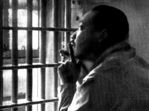 Martin Luther King, Jr. sitting with his hand to his chin, looking through the bars of a jail cell