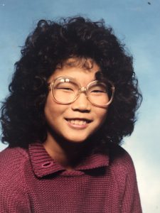 Rebecca Slaby's yearbook photo from the 7th grade, featuring her Meg Ryan-inspired perm. She is smiling and wearing large, clear glasses and a pink sweater.