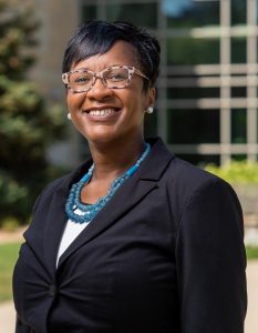 Dr. Tyner smiling wearing glasses, a black blazer, a white shirt, and a long blue necklace.