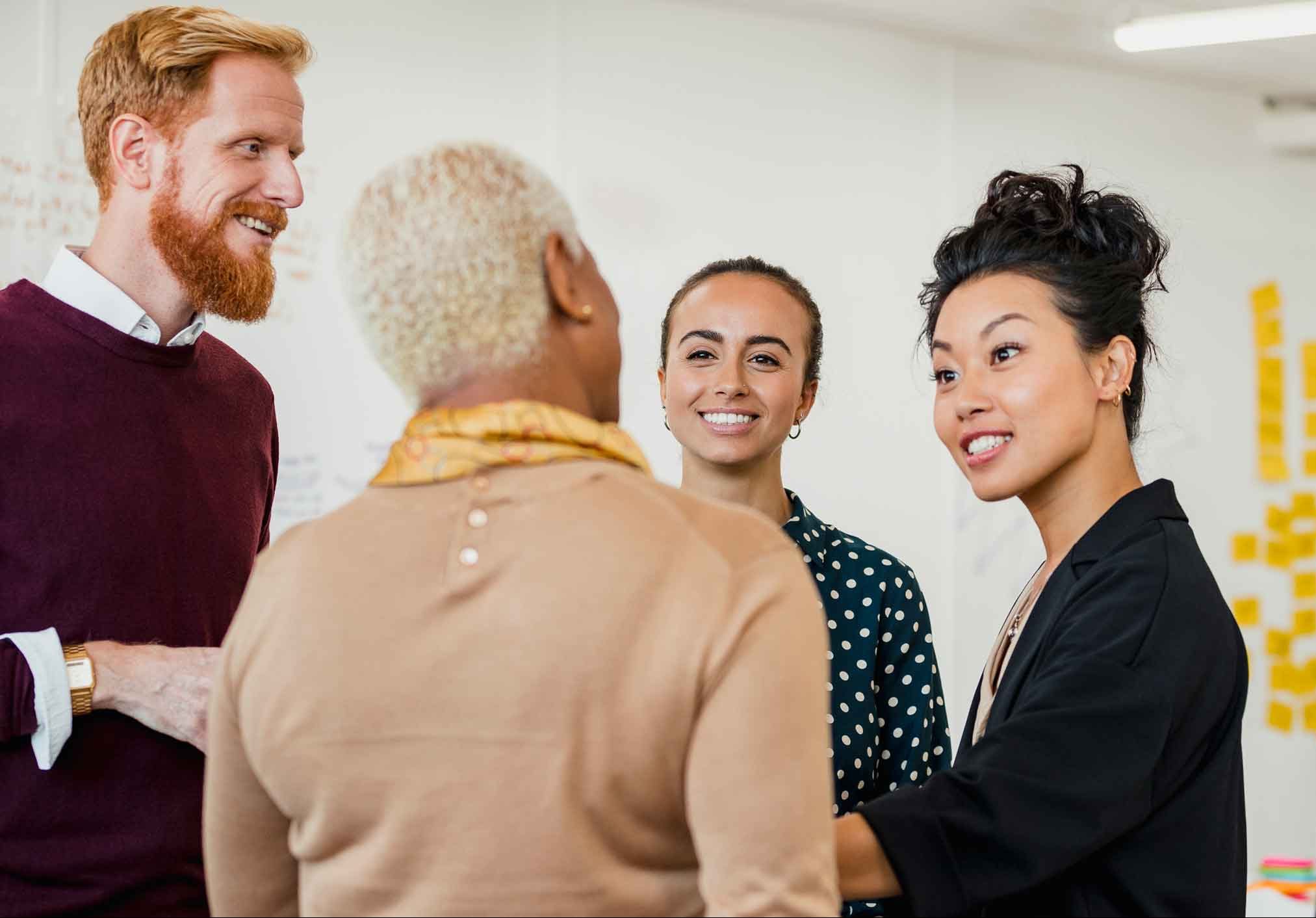 Four diverse coworkers of various ages engage in conversation in the office, with post-its on the back wall