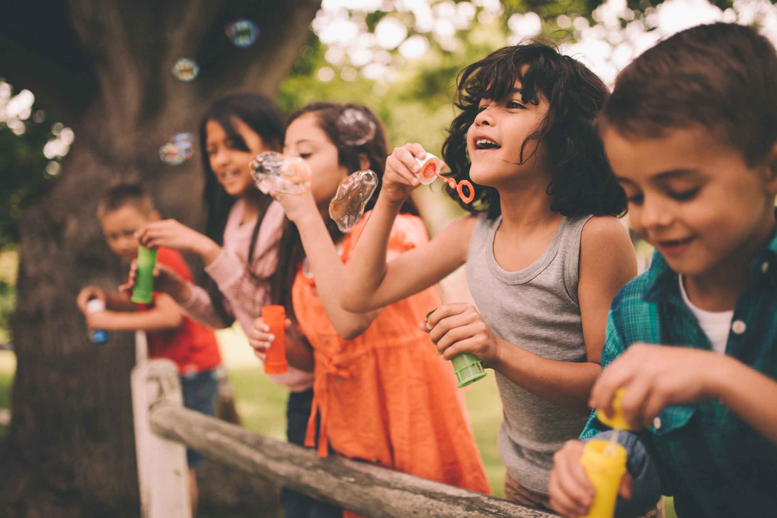 5 kids leaning up against a fence, blowing bubbles and smiling