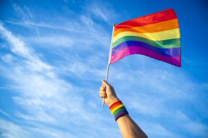 A hand wearing a rainbow wristband waves a pride flag in the air with a cloudy blue sky in the background
