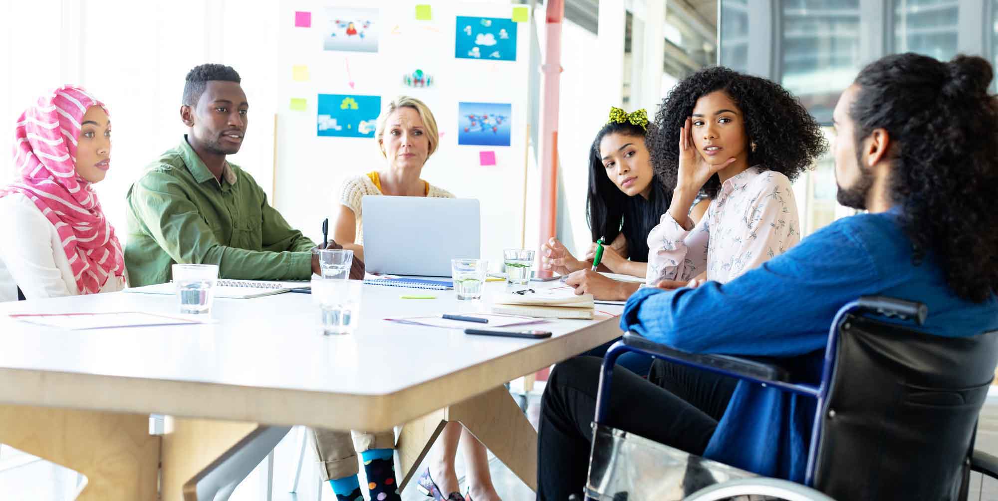 A group of six diverse coworkers engaged in conversation around a conference table in a brightly lit, windowed office