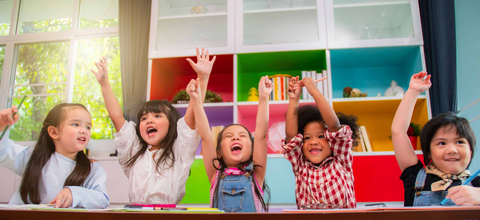 Six young children in bright clothing smile and raise their hands in a brightly colored, sunlit room