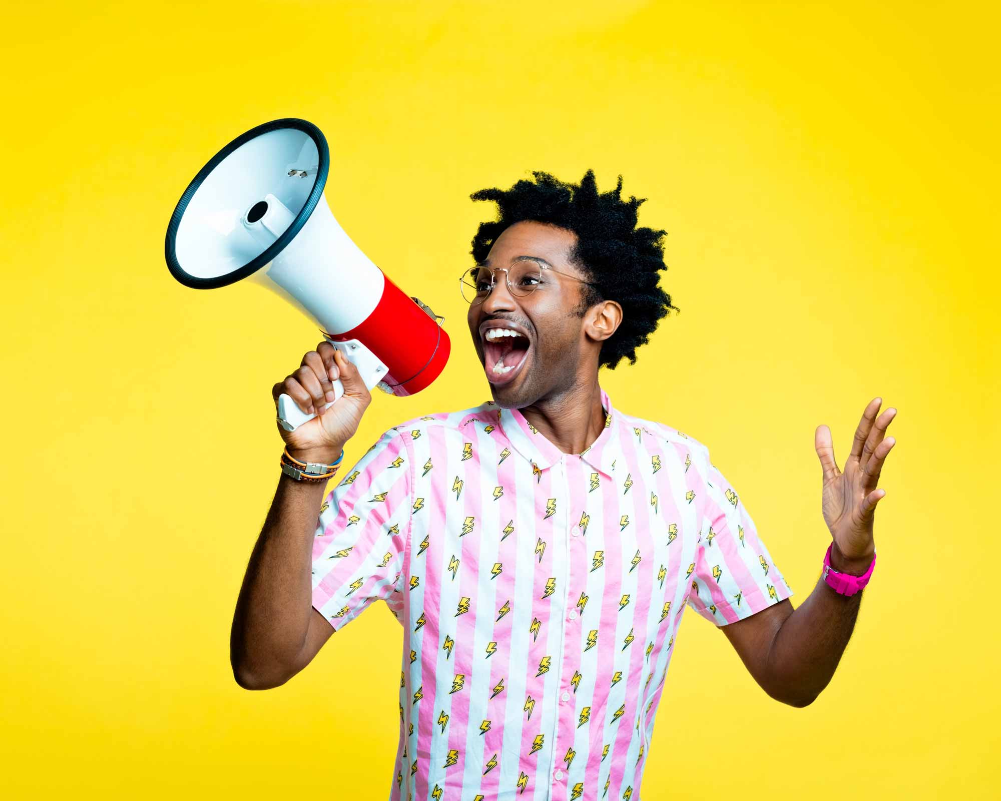 Man in a red patterned shirt smiling and speaking into a megaphone against a bright yellow background