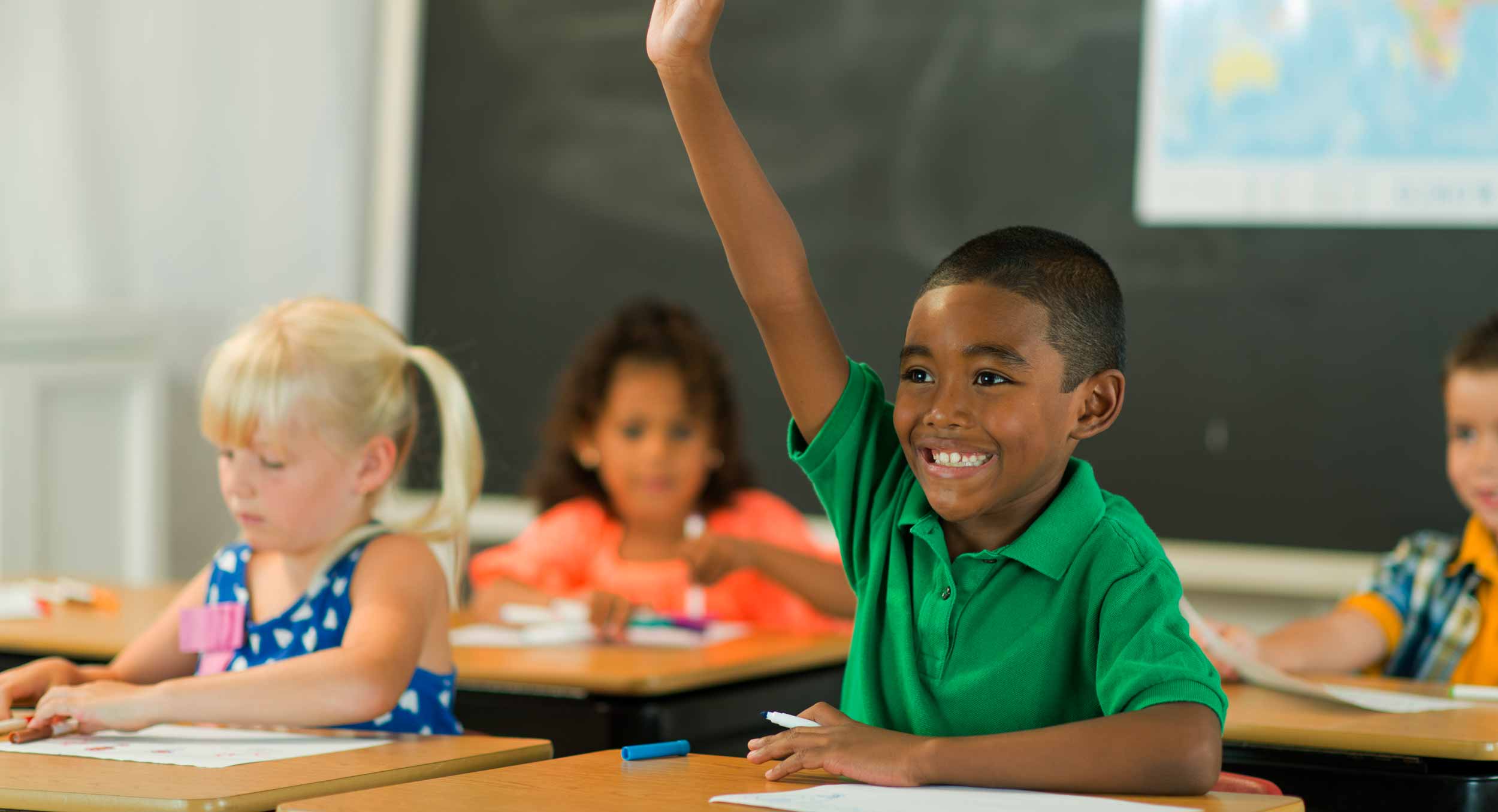 A smiling boy in a green polo raises his hand in an elementary school classroom