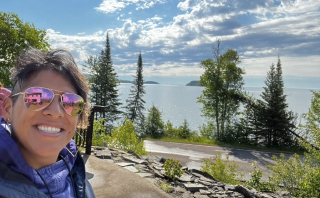 Juana standing in front of a scenic background of a lake and trees, smiling and wearing sunglasses