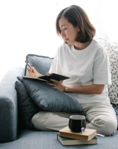 A person sitting on a comfy couch and journaling, with a cup of tea and a book next to her