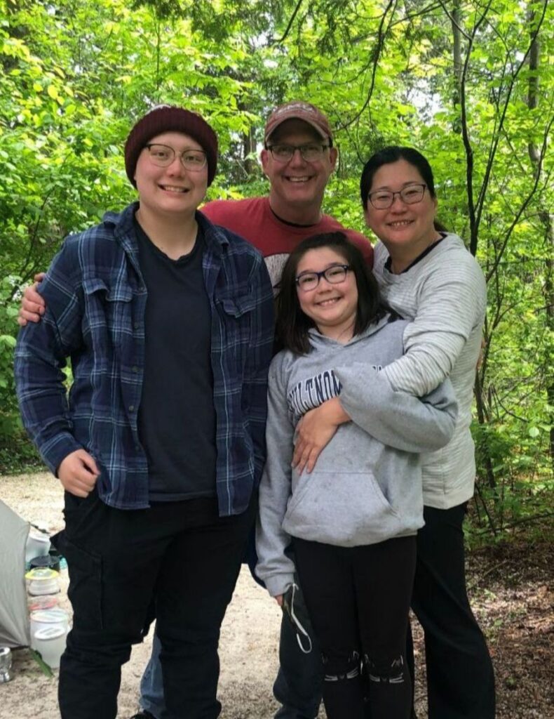 Executive Director, Rebecca Slaby, hugging and standing with her husband, Colin, and two children, Emma and Clara. They are surrounded by beautiful green trees.