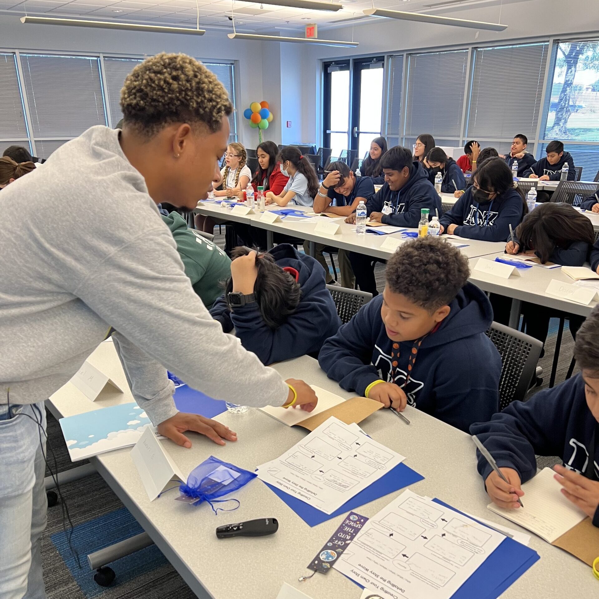 Franklin the Helper leading a book writing workshop. Rows of students sitting and writing at tables. Franklin is leaning over to assist one student.