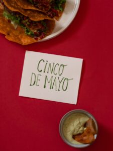 Namecard reading, "Cinco de Mayo" on a table with a red tablecloth, featuring two tacos on a plate