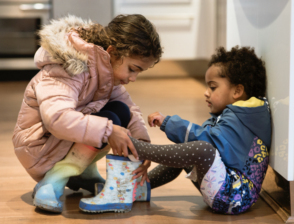 A child helping her younger sibling put on a rain boot
