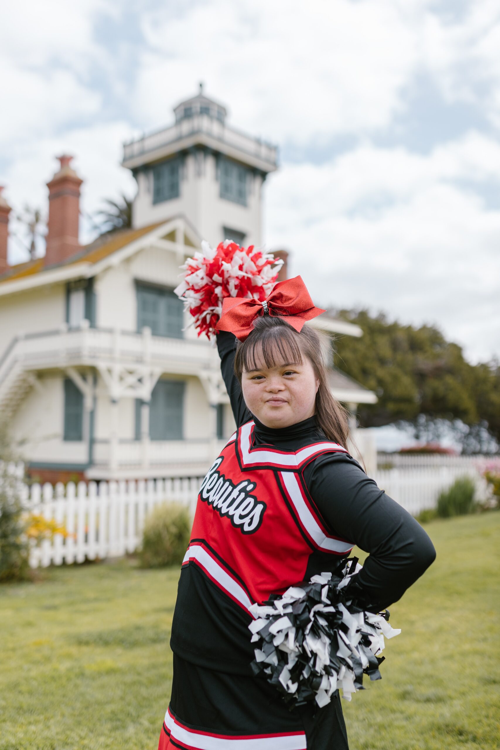 a cheerleader with down syndrome wearing a black, red, and white uniform. She is holding one black and white pom pom on her hip and one red and white pom pom above her head. She stands outside in the grass in front of a large house and cloudy sky.