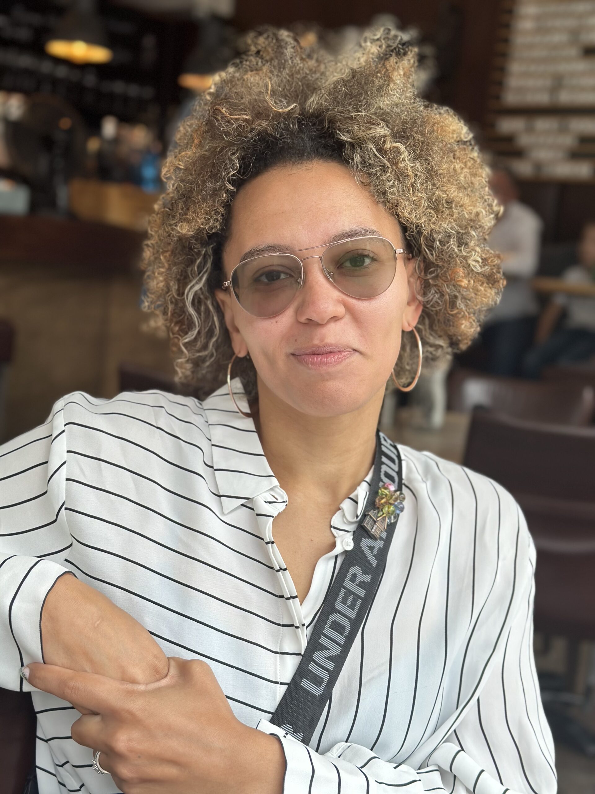 Photo of Sydney Carlson, a mixed race (Black and White) woman wearing a white button up shirt with horizontal and vertical black stripes. She has blonde curly hair and is wearing sunglasses and hoop earrings.