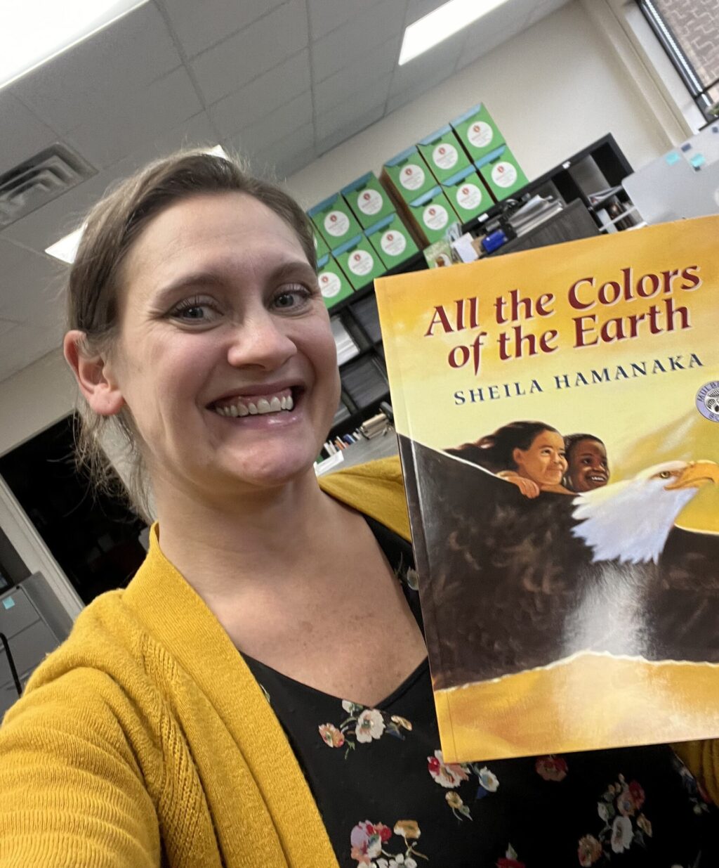 A selfie of Jenni posing with children's book, "All the Colors of the Earth." The book pictures two children riding a flying eagle in a yellow sky. Jenni wears a matching yellow cardigan and a black floral shirt.