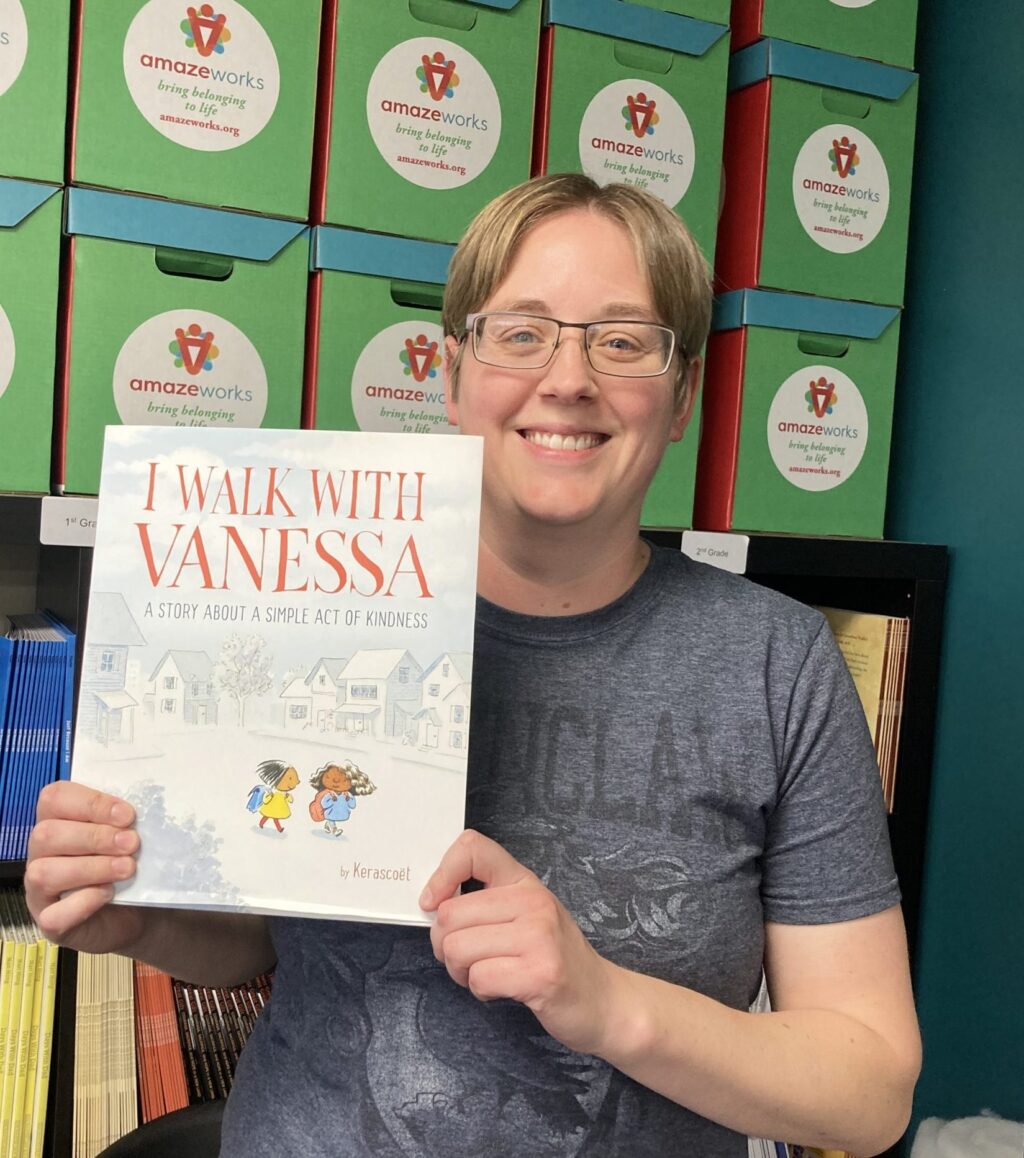 Melissa Hendrickx smiling and posing with the picture book, "I Walk with Vanessa." She is wearing a gray t-shirt and standing in front of an AmazeWorks book shelf with book boxes stacked on top of it.