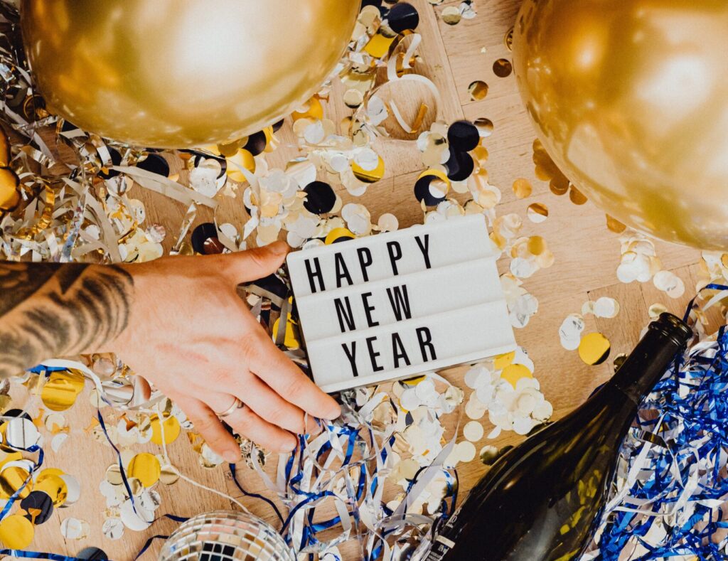 A sign reading, "Happy New Year," atop of holiday decorations, including golden balloons, confetti, and a champagne bottle