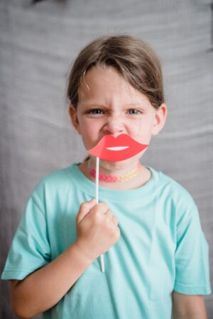A child frowning while holding prop cartoon lips to their mouth, maybe being forced to engage in toxic holiday joy!
