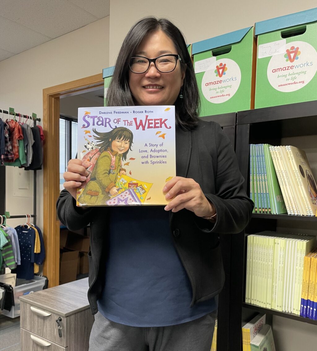 Rebecca posing with the picture book, "Star of the Week," and smiling. She is standing in front of an AmazeWorks book shelf with book boxes stacked on top.