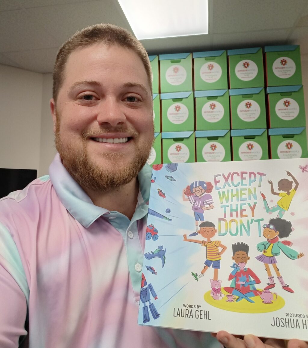 A selfie of Ryan posing with picture book, "Except When They Don't." He is wearing a pink pastel tie dye collared shirt, and AmazeWorks book boxes are stacked behind him. 