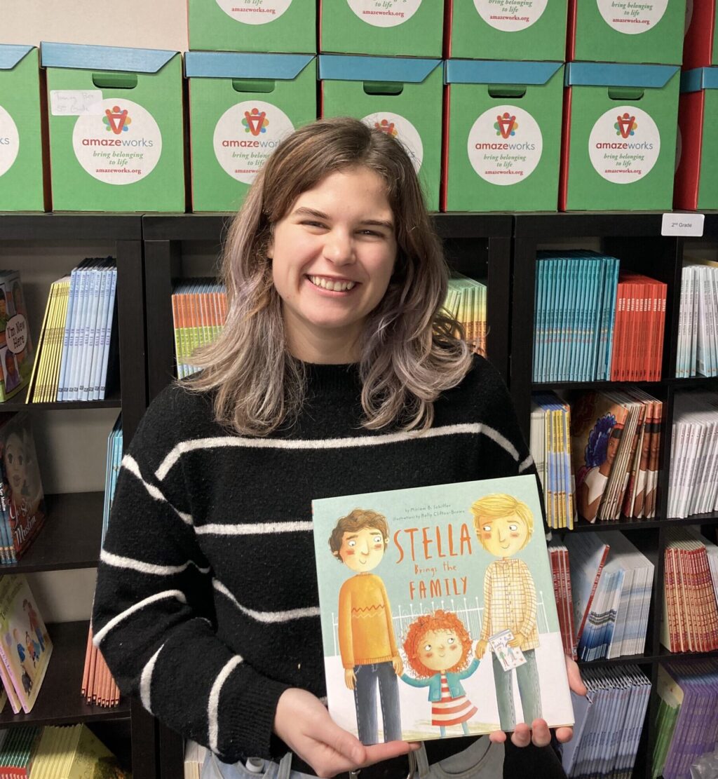 Sophie holding the picture book, "Stella Brings the Family," featuring a red-headed child holding hands with her two dads. Sophie is wearing a black sweater with thin white stripes and is standing in front of the AmazeWorks book shelf with book boxes stacked on top.