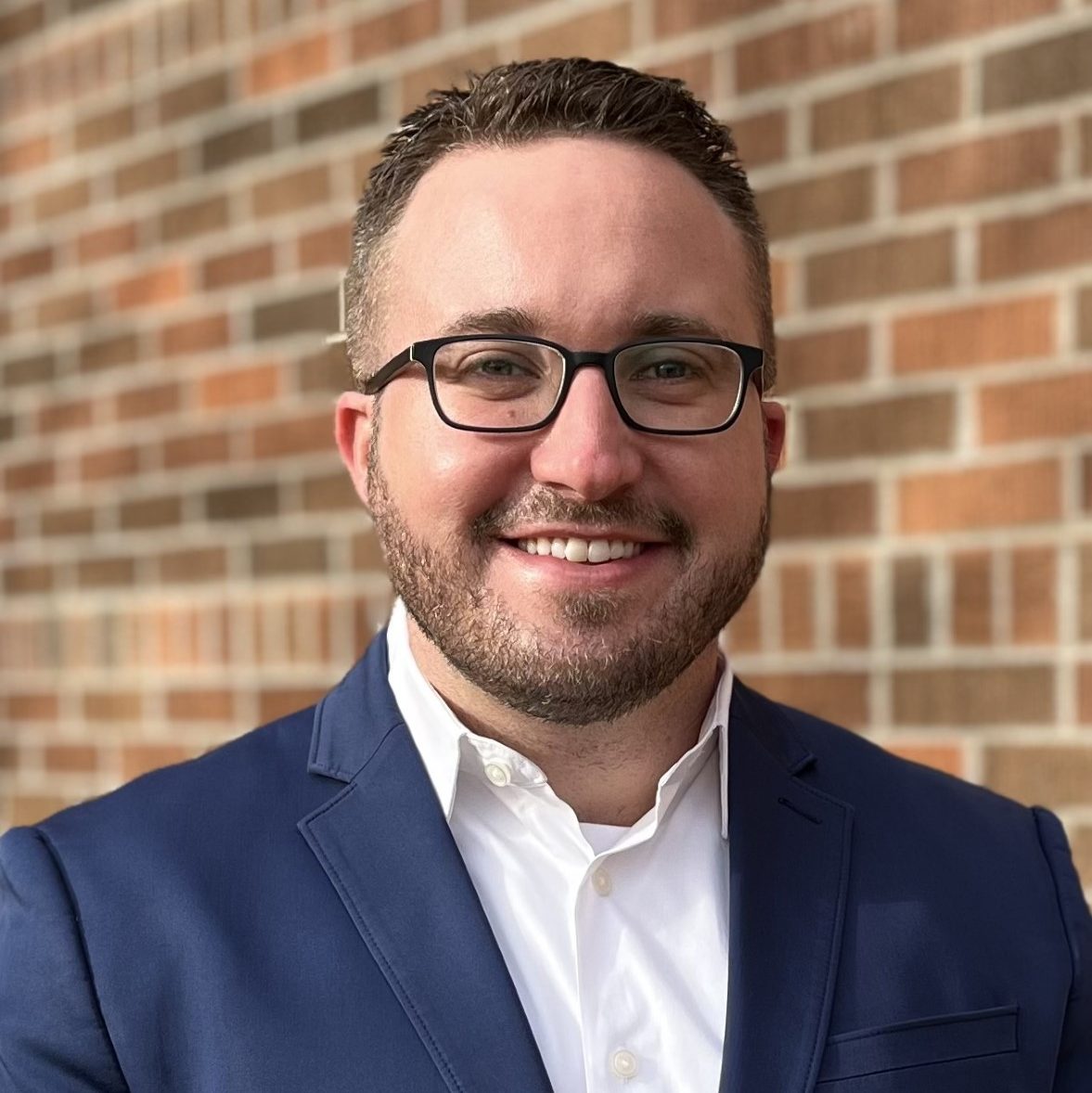 Headshot of John Bjoraker wearing a navy blue blazer, white button collared button up, and black glasses. He is smiling and standing in front of a brick wall.