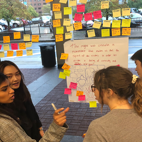 Three college students doing group work. They have a large poster with writing surrounded by orange, yellow, and pink sticky notes.