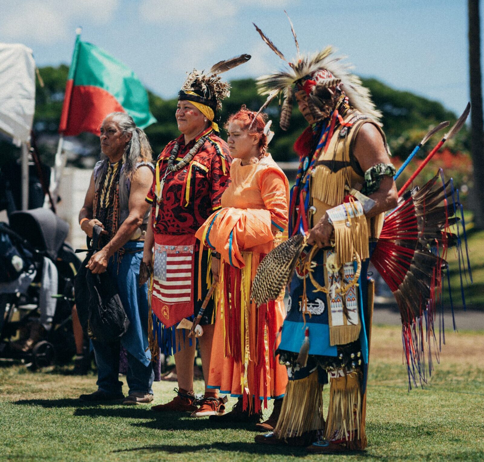 Four Native people wearing traditional Native dress, including headdresses and moccasins, at an outdoor event.