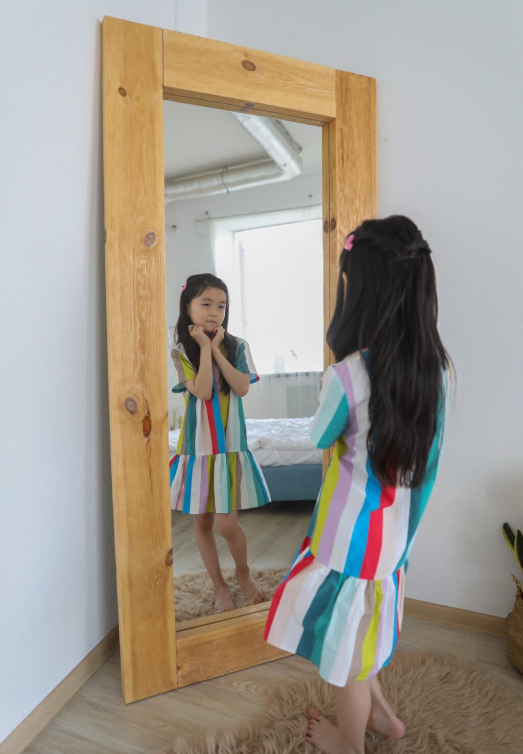 A child wearing a colorful dress with vertical stripes looking into a full length mirror with a wooden frame leaned against the wall. She poses with her hands curled into fists under her chin and her right knee bent inward.