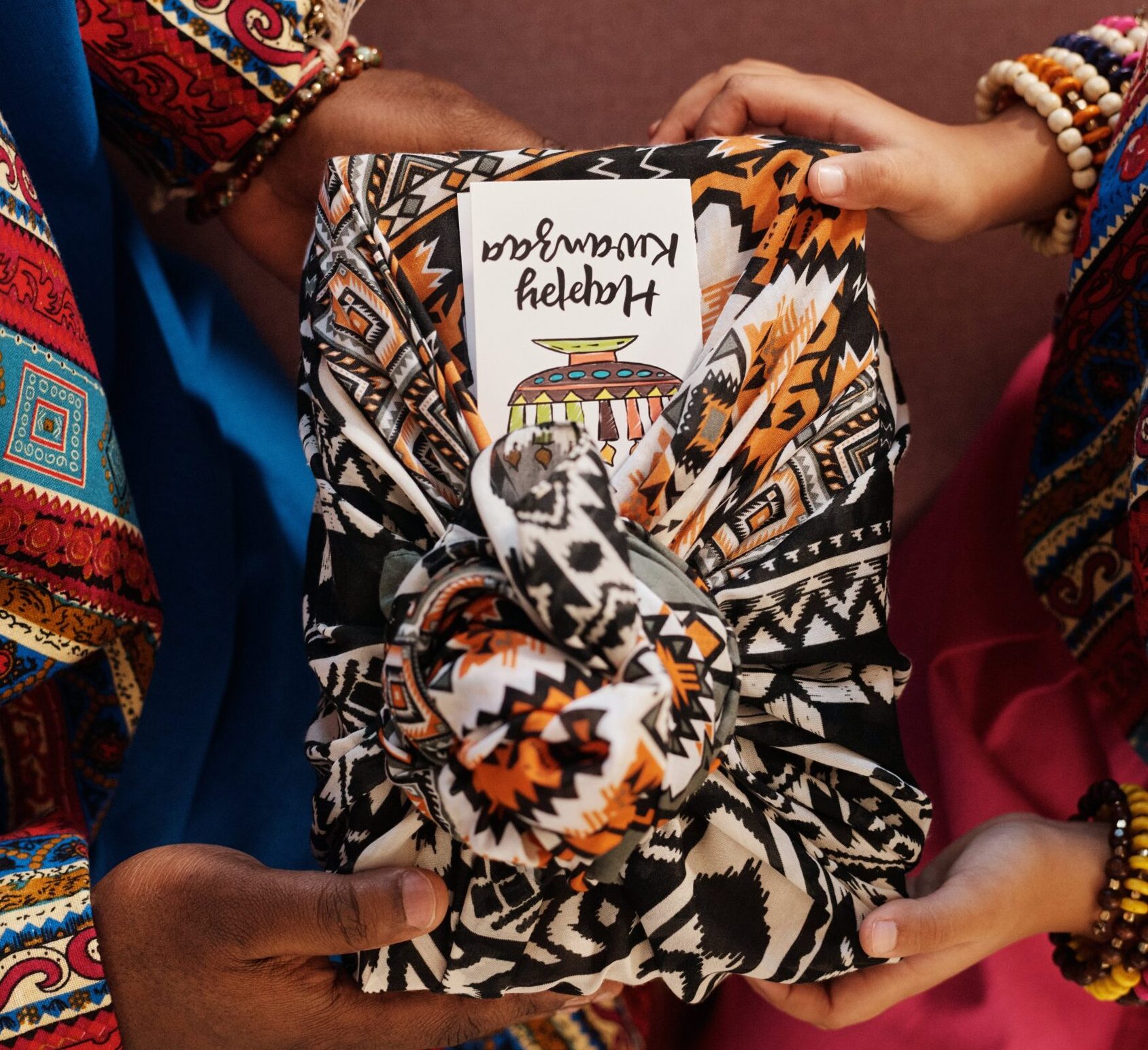Two pairs of hands holding a gift wrapped colorfully with a note reading, "Happy Kwanzaa"