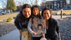 Two caregivers smiling on either side of their child who holds a copy of The Rock in My Throat