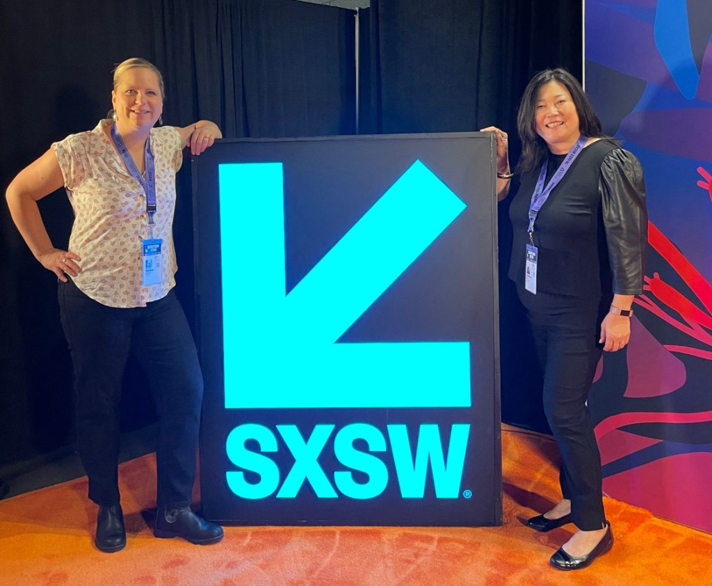 Mel Andersen and Rebecca Slaby stand and smile with their hands resting on either side of a neon blue SXSW sign
