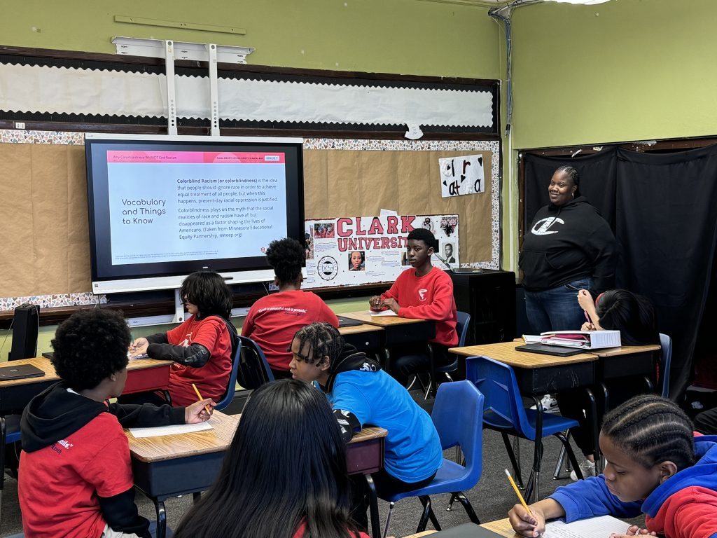 Evelyn Stevenson's middle school classroom during an AmazeWorks lesson, Why Colorblindness Will NOT End Racism." Students wearing red sweatshirts are seated at their desks listening to Evelyn, who stands at the front of the room.