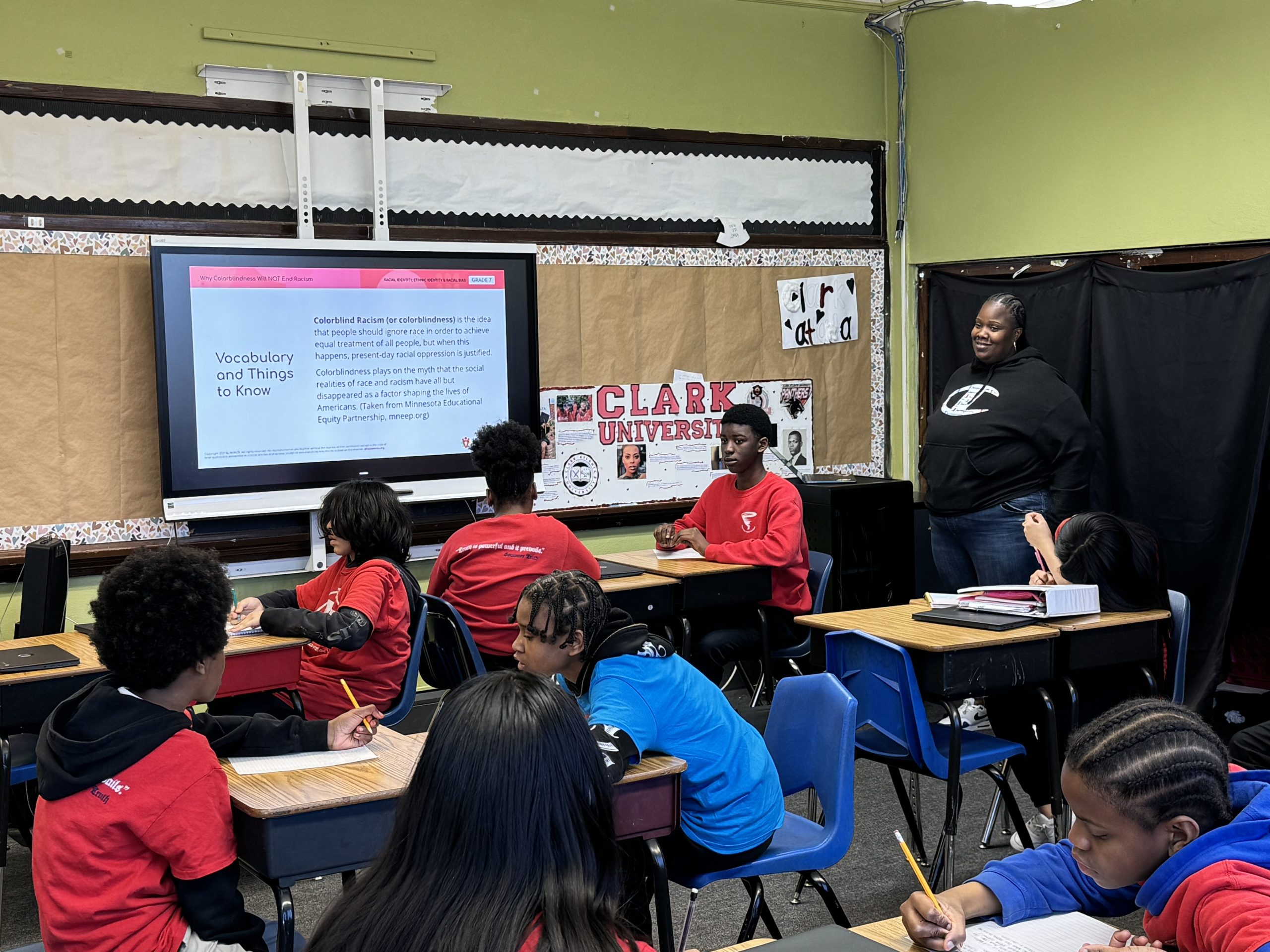 Evelyn Stevenson's 6th grade classroom during an AmazeWorks lesson, Why Colorblindness Will NOT End Racism." Students wearing red sweatshirts are seated at their desks listening to Evelyn, who stands at the front of the room.