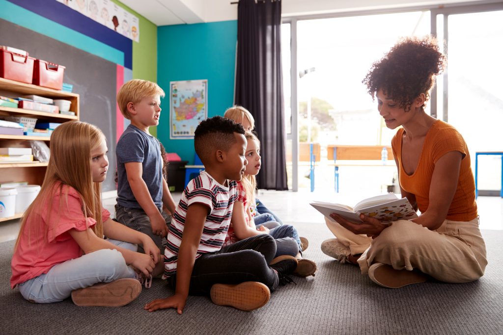 A teacher sitting on the floor holding a book, reading aloud to five students.