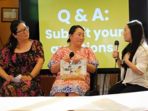 Rebecca Slaby, Kao Kalia Yang, and Khou Vue sit on a stage. Khou speaks into a microphone as Rebecca and Kalia listen. Kalia holds a copy of Caged in her lap.