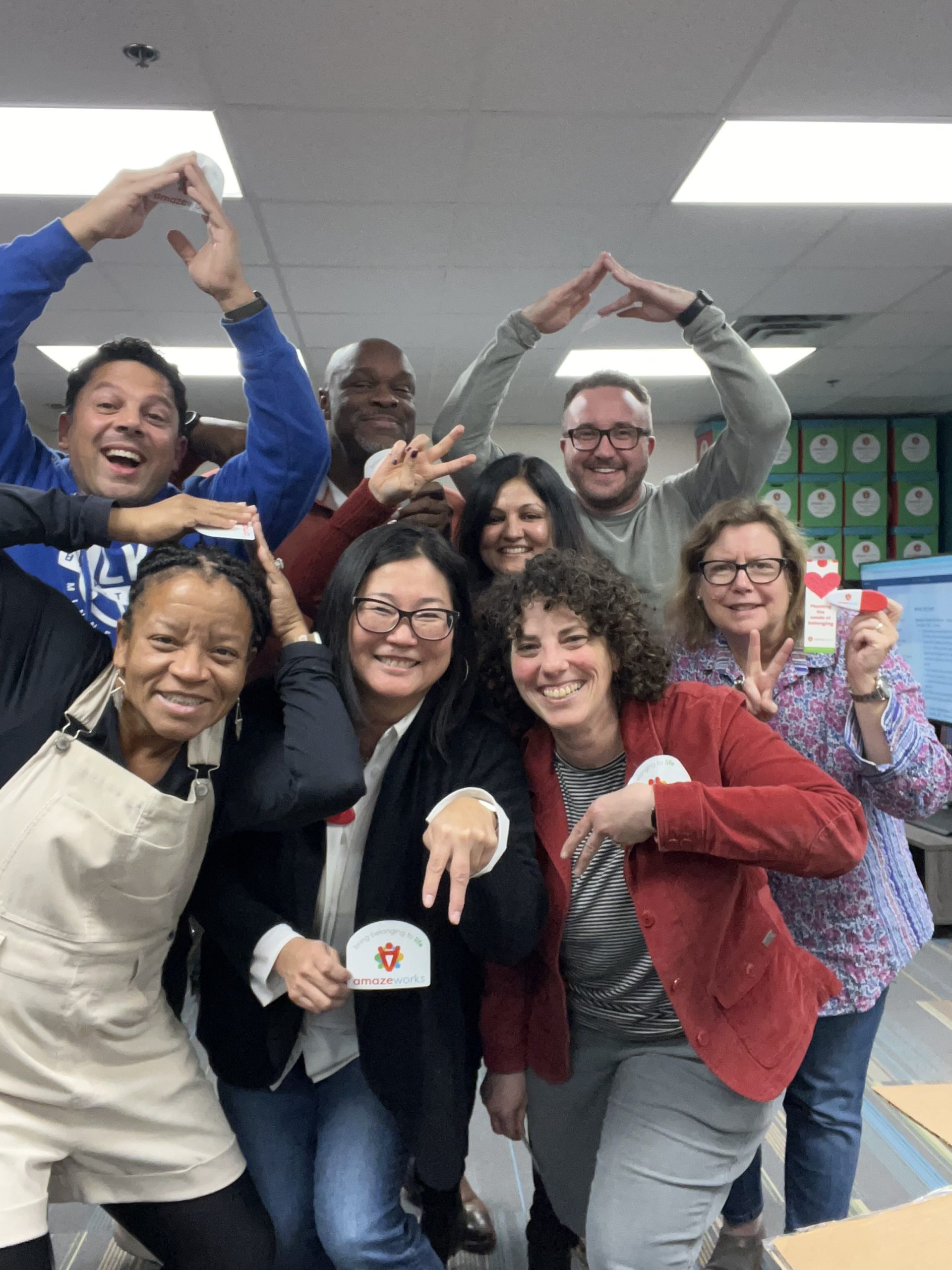 AmazeWorks board smiling and creating the letter "A" with their hands. They hold up AmazeWorks stickers and bookmarks.