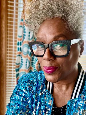 A selfie of Timi Bliss. Timi is a Black woman with curly gray hair. She is wearing black glasses, pink lipstick, and a blue sequin jacket.