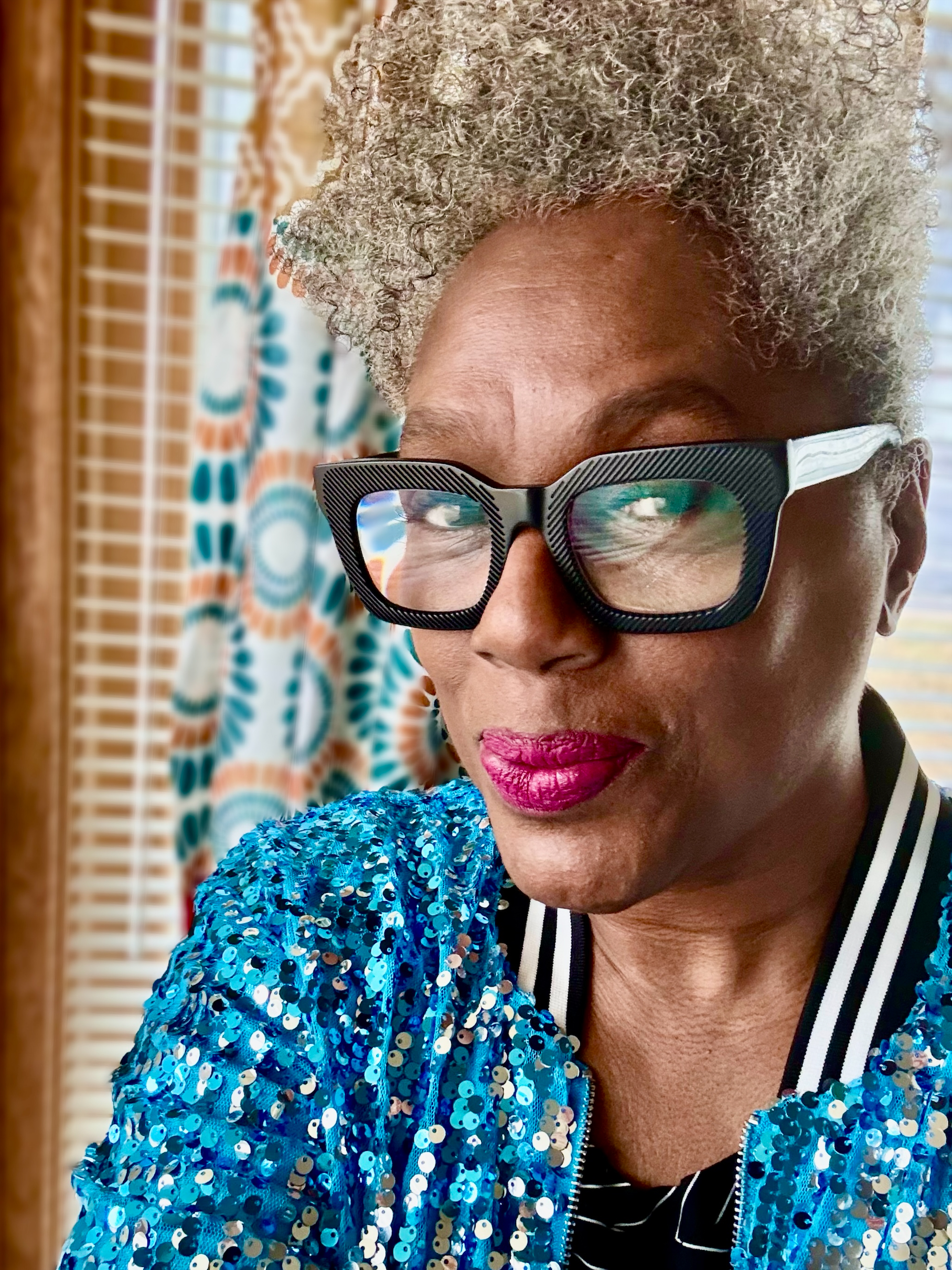 A selfie of Timi Bliss. Timi is a Black woman with curly gray hair. She is wearing black glasses, pink lipstick, and a blue sequin jacket.