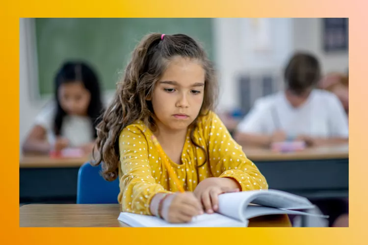 A young child with long, curly brown hair wearing a yellow long-sleeve shirt. She writes with a pencil in a notebook, sitting at a desk in her classroom.