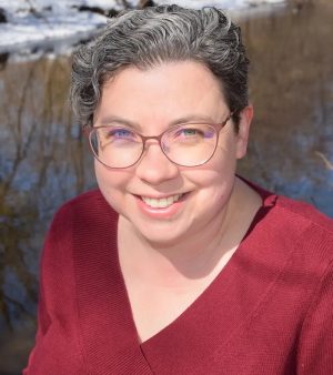 Smiling headshot of Molly Beth Griffin, a White woman with short curly gray hair, rose gold glasses, and a red v-neck sweater.