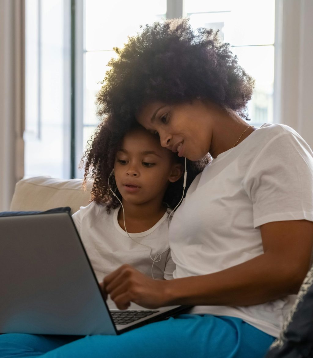 A Black woman with an afro and white t-shirt sits with her child with medium-length curly hair. They're wearing earphones and watching something together on a laptop and look mid-conversation.