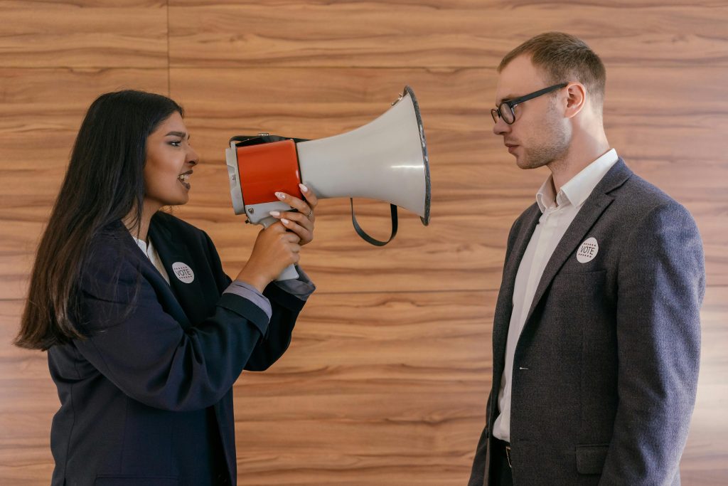 A BIPOC woman with long brown hair wearing a suit holds a megaphone to her mouth, facing a White man also wearing a suit, making a displeased face.