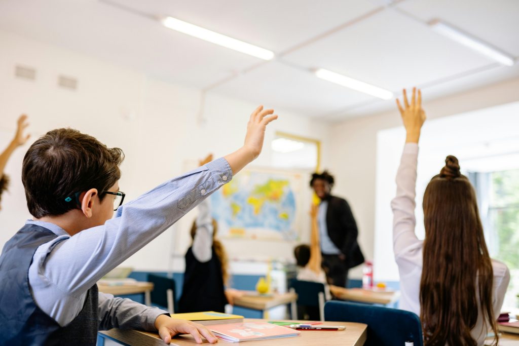 Children raise their hands in a classroom, waiting for their teacher to call their name