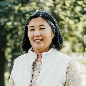 Headshot of Kari Scanlon, a Korean woman with shoulder-length black and gray hair. She is smiling and wearing an open white vest over a patterned shirt.