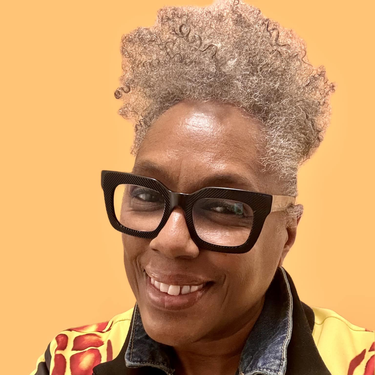 Headshot of Timi Bliss, a Black woman with curly gray hair. Timi is smiling and wearing black glases and a yellow and black jacket.