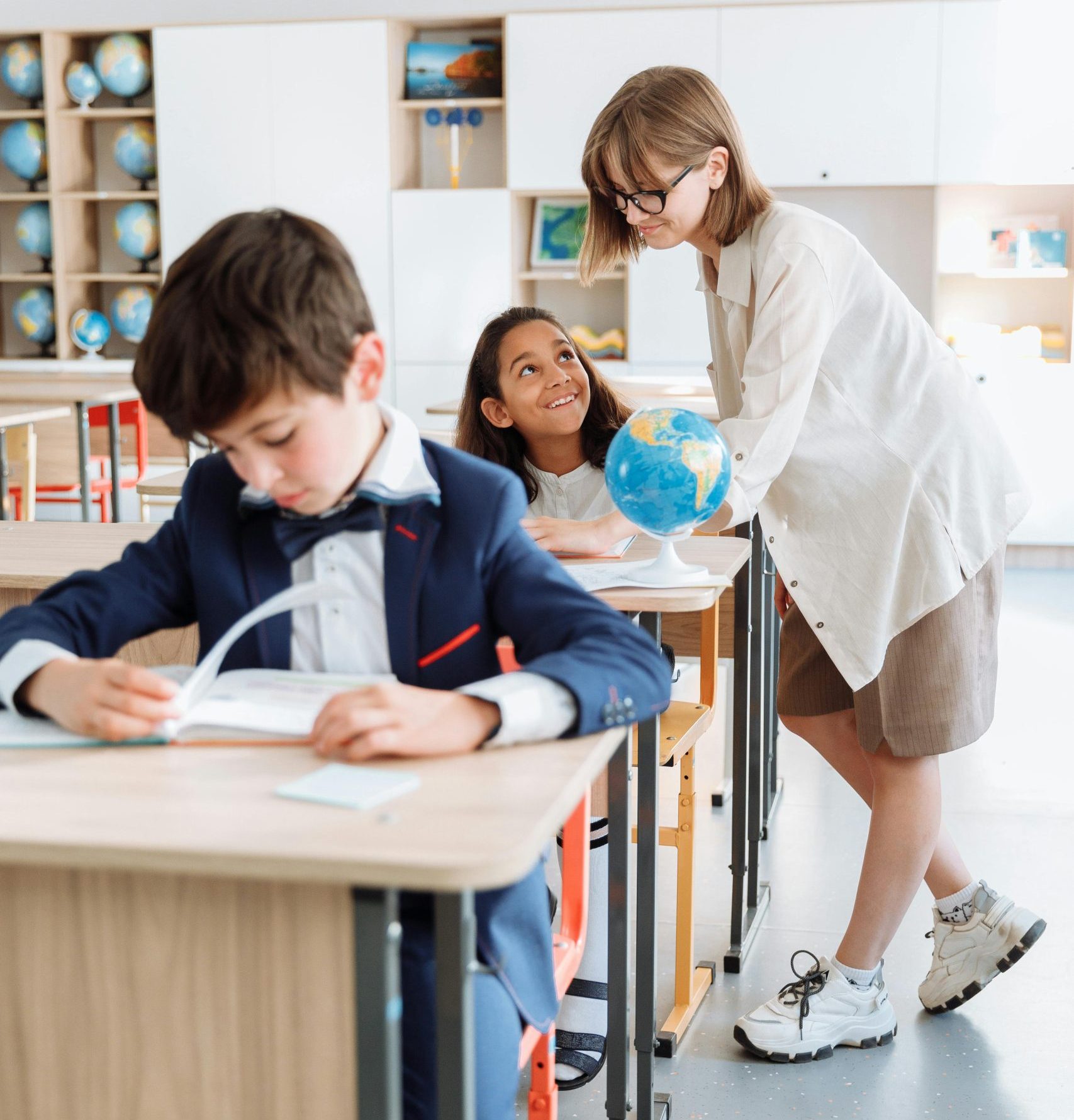 Teacher stands next to a student's desk to help her. Another student sits in his desk in the foreground.