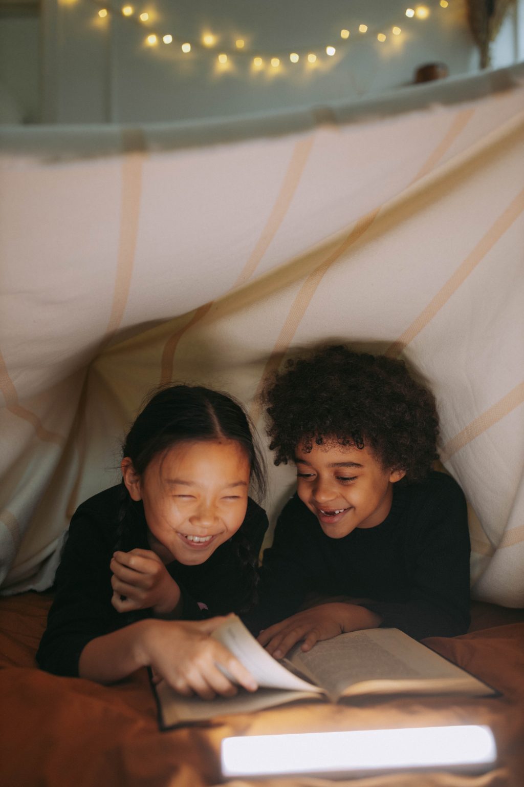 An Asian girl and a Black boy giggle together while reading a book in a blanket fort.