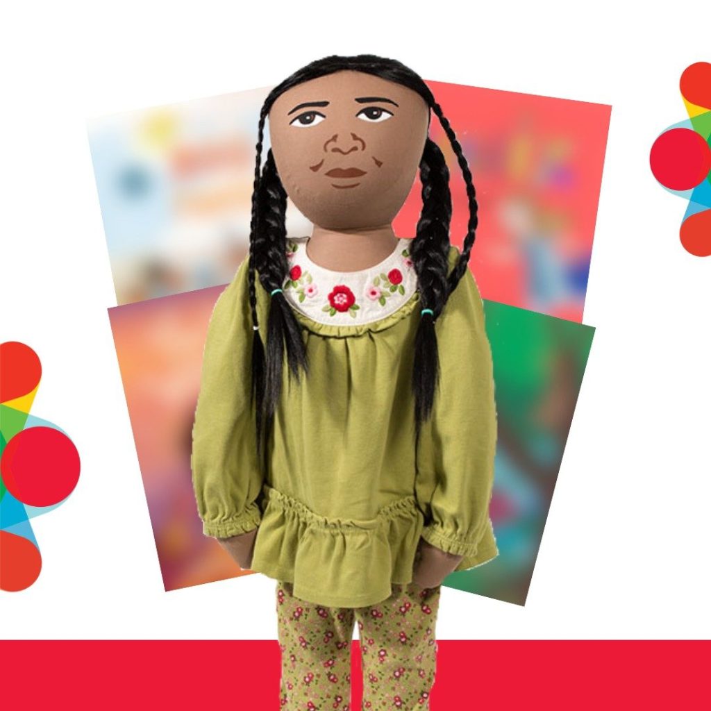 Leela the Persona Doll stands in front of four blurred picture book covers. Leela is wearing a green floral shirt and pants with braided hair.